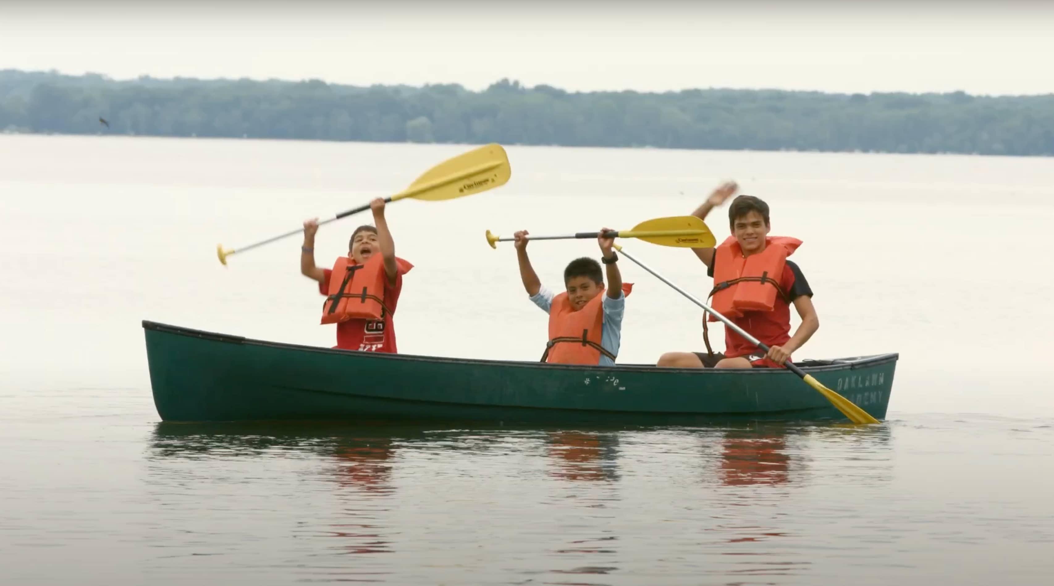 Cuatro estudiantes en chaleco salvavidas remar en bote verde con paletas amarillas en lago tranquilo.